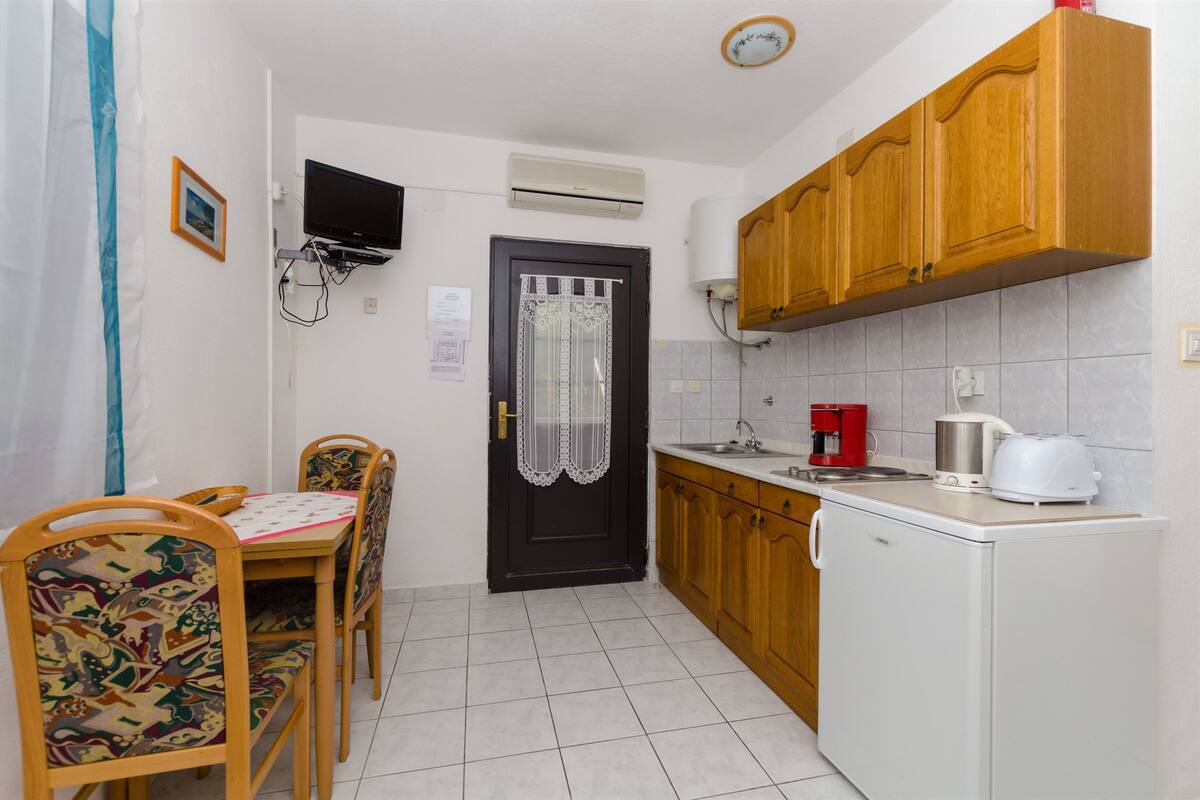A compact kitchen with a dining table, chairs, a refrigerator, a coffee maker, and a TV mounted on the wall.