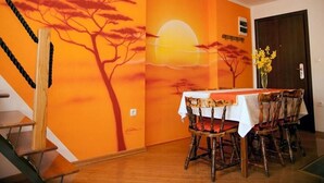 A dining area with a table set for four, surrounded by wooden chairs with red cushions. A mural of a sunset with trees is on the wall behind the table. - Apartment Sunset (Vodice)