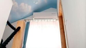 A room with a sloped ceiling painted to resemble a sky with clouds, a window with pink curtains, and an air conditioning unit mounted on the wall. - Apartment Sunset (Vodice)