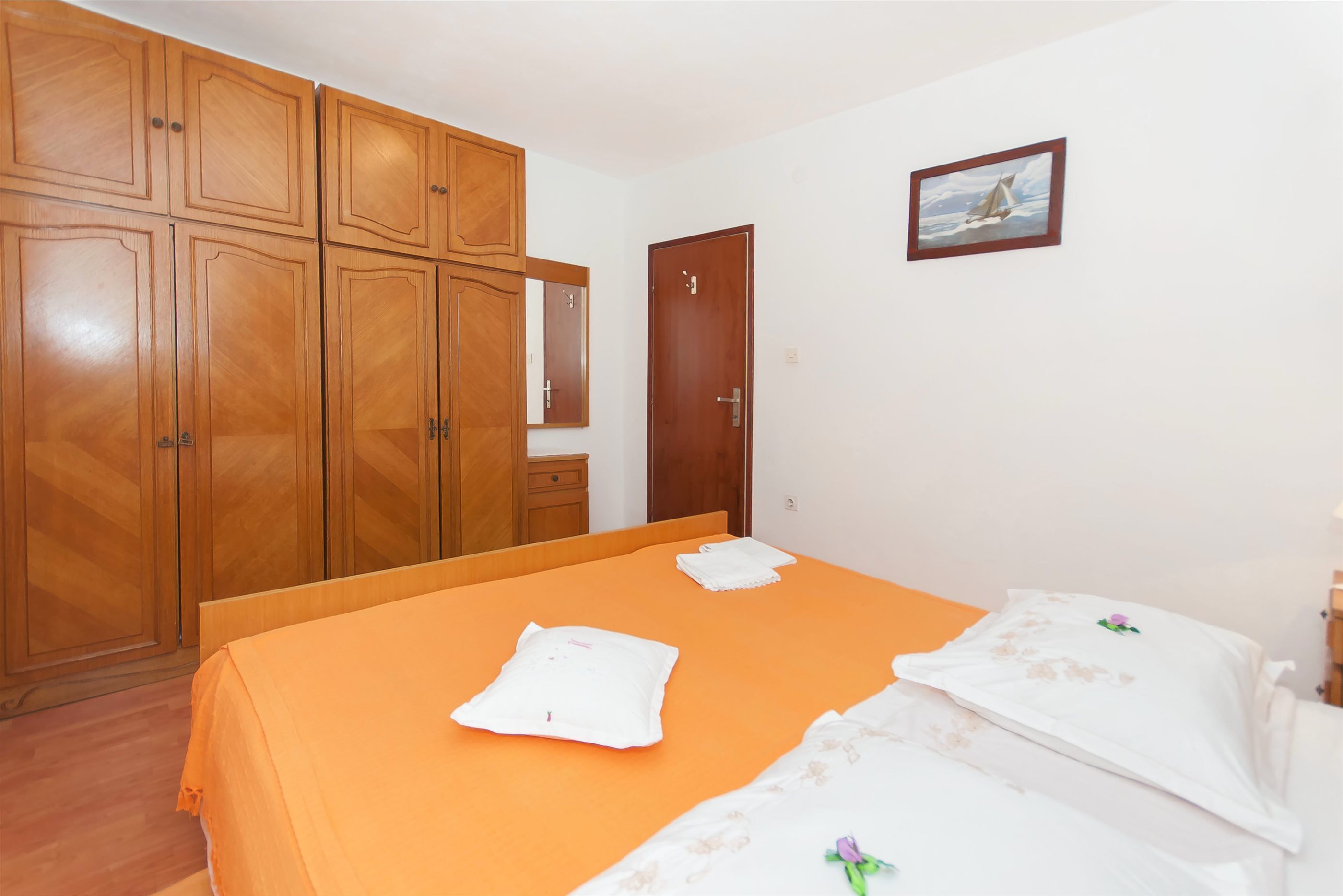 A bedroom with a wooden wardrobe, a bed with an orange bedspread, and a door with a small picture on the wall.