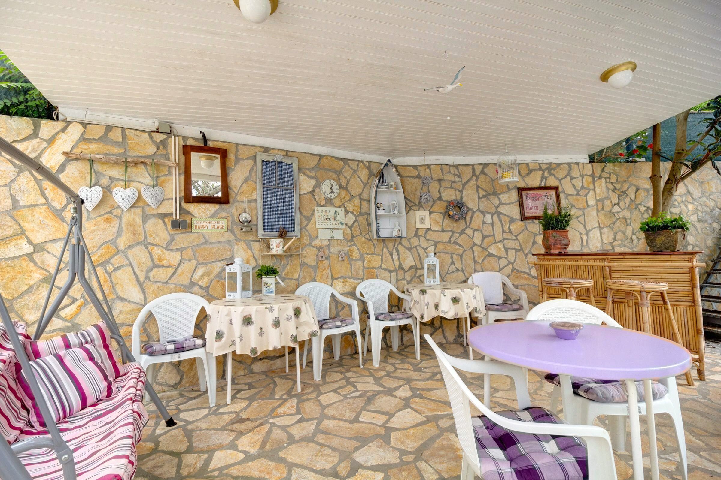 Eine Terrasse mit Steinwänden, weißer Möbel, einem Schaukelstuhl und einem Tisch mit lila Tischdecke.