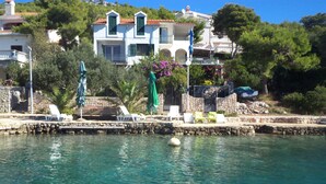 Une propriété située sur la côte, dotée d’un mur en pierre, d’une eau d’un bleu limpide et d’un bâtiment blanc aux volets verts.