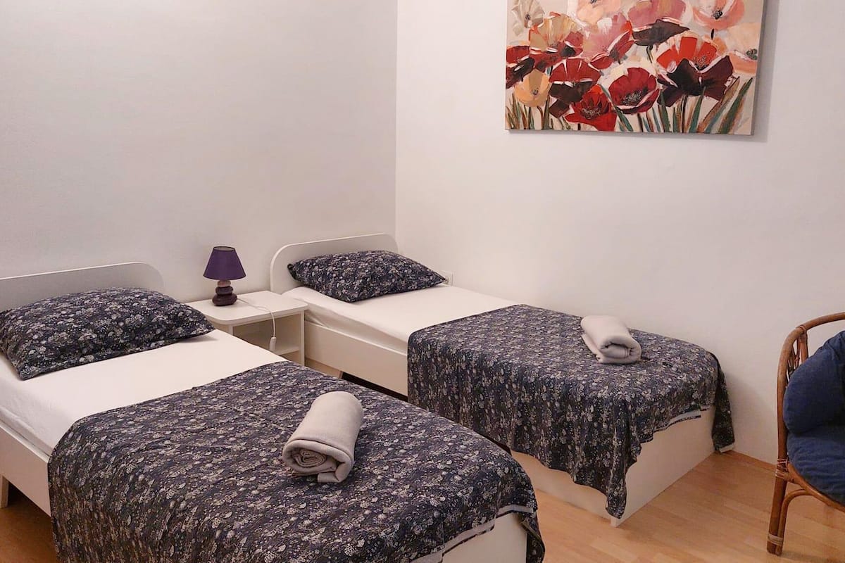 Ein Zimmer mit zwei Betten, jeweils mit floralem Überwurf und einem zusammengerollten Handtuch. Ein Sessel, ein Nachttisch mit Lampe und ein Bild an der Wand sind ebenfalls vorhanden.