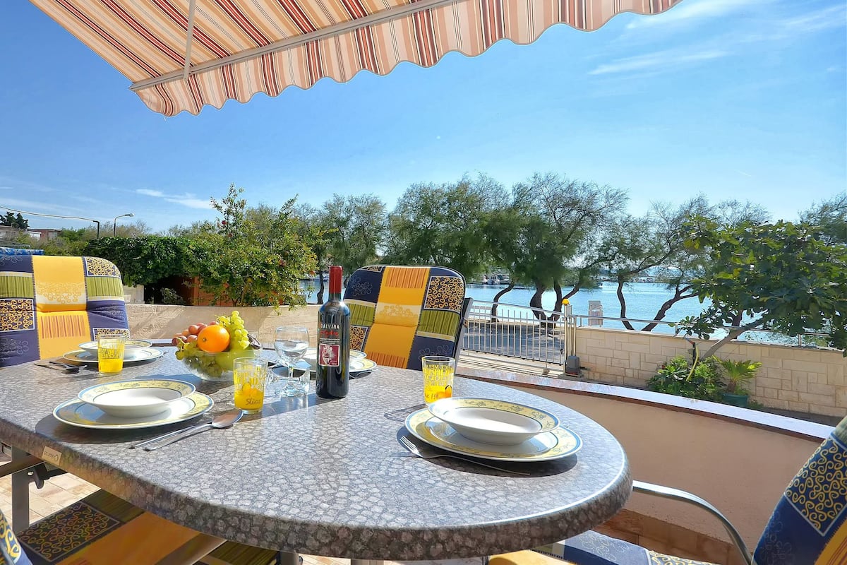 An outdoor dining area with a table set for two, featuring patterned chairs and a view of the sea.