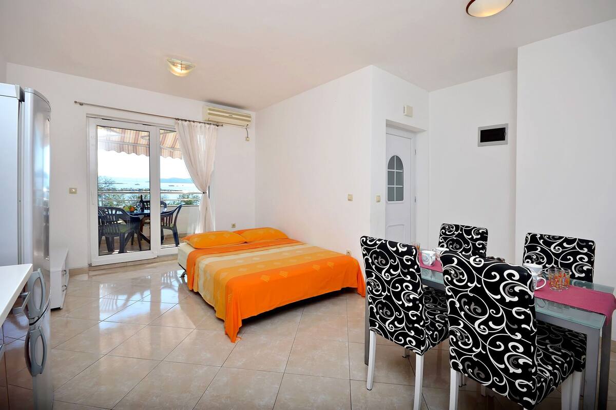 A hotel room with a bed, a dining table with chairs, a washing machine, and a view of the sea through the window.