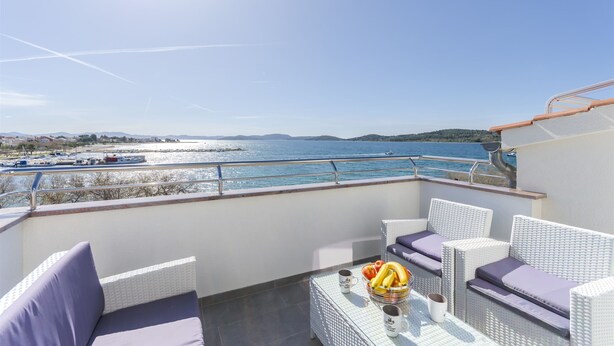 Un balcón con mobiliario de mimbre, una mesa con frutas y bebidas, y vistas al mar y barcos.
