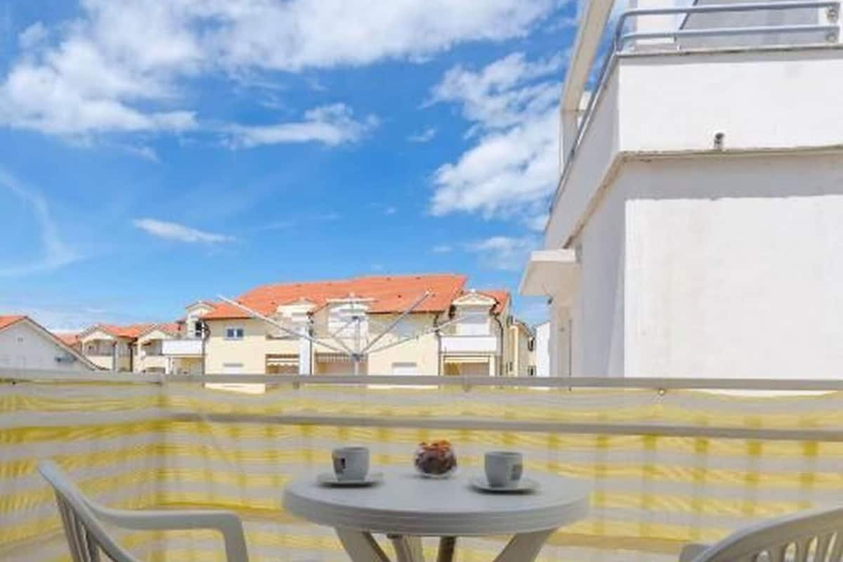 A balcony with a yellow checkered tablecloth, two white plastic chairs, and a small table with cups and a flower.