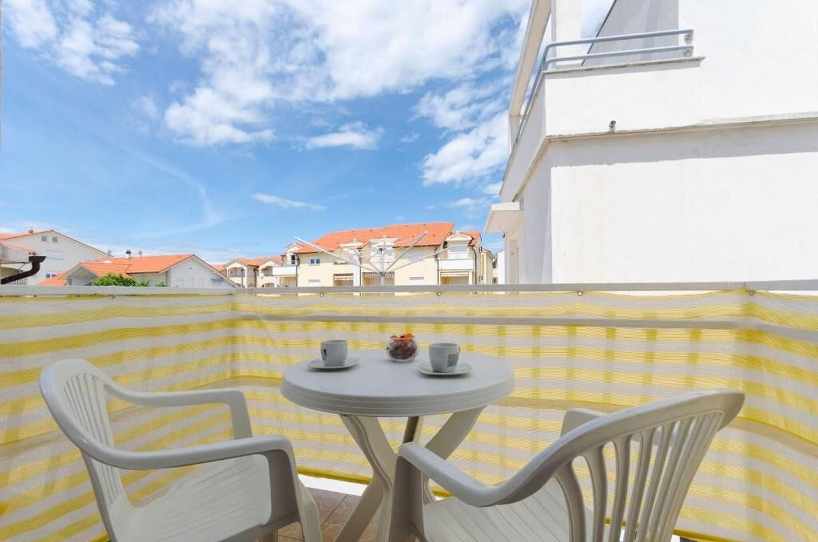 A balcony with a yellow striped partition, two white plastic chairs, and a small round table with two cups and a small vase.