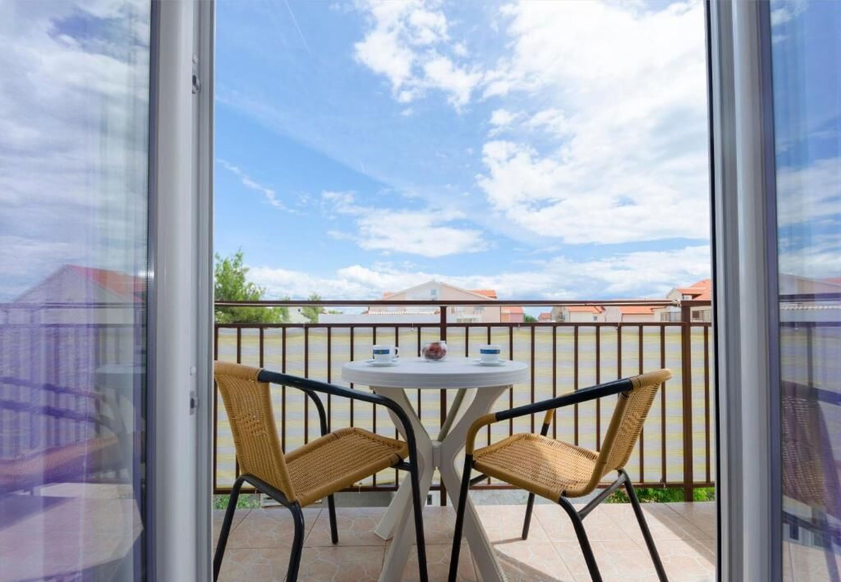 A balcony with two wicker chairs and a small round table, overlooking a fenced area with buildings in the background.