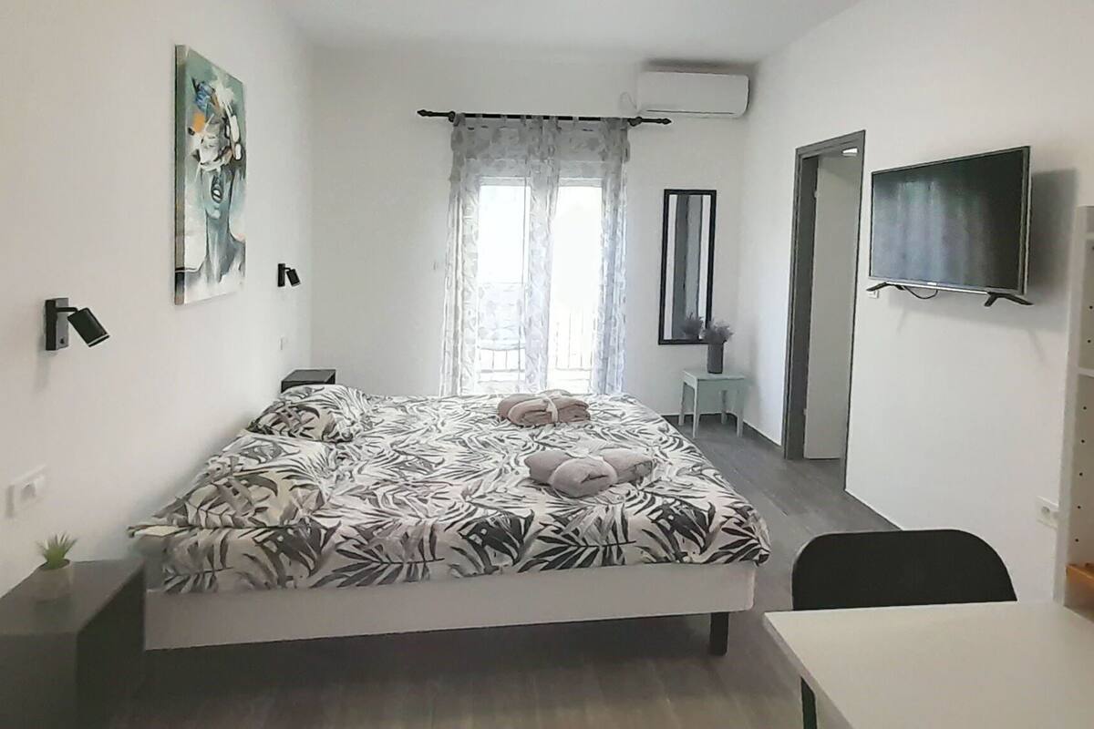 A bedroom with a bed, a desk, a chair, a TV, a wardrobe, and a window with curtains.