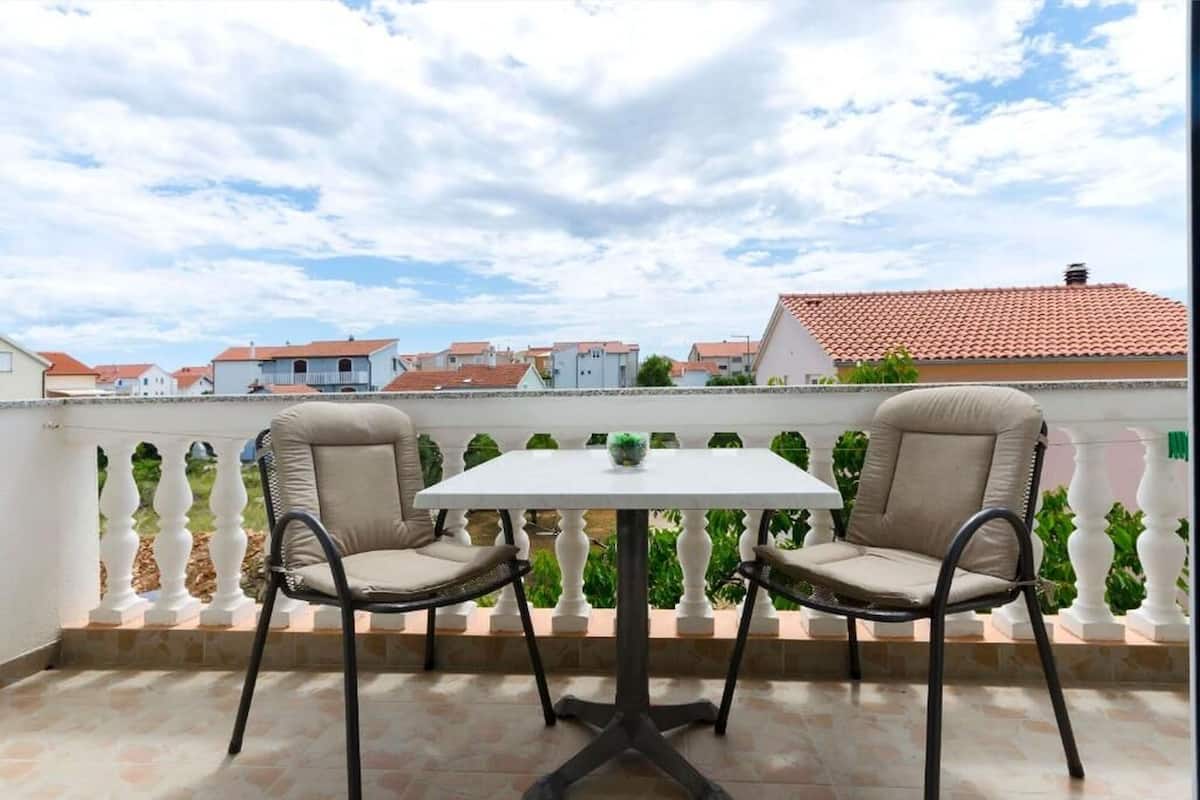 A balcony with two chairs and a table, overlooking a residential area with red-tiled roofs.