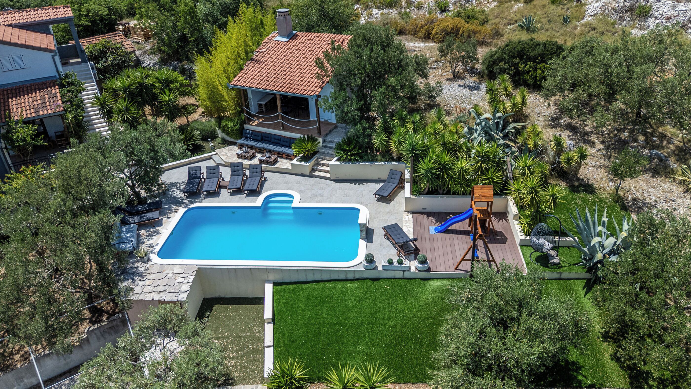 An aerial view of a residential property with a swimming pool, sun loungers, a children's play area, and a small house with a red-tiled roof.