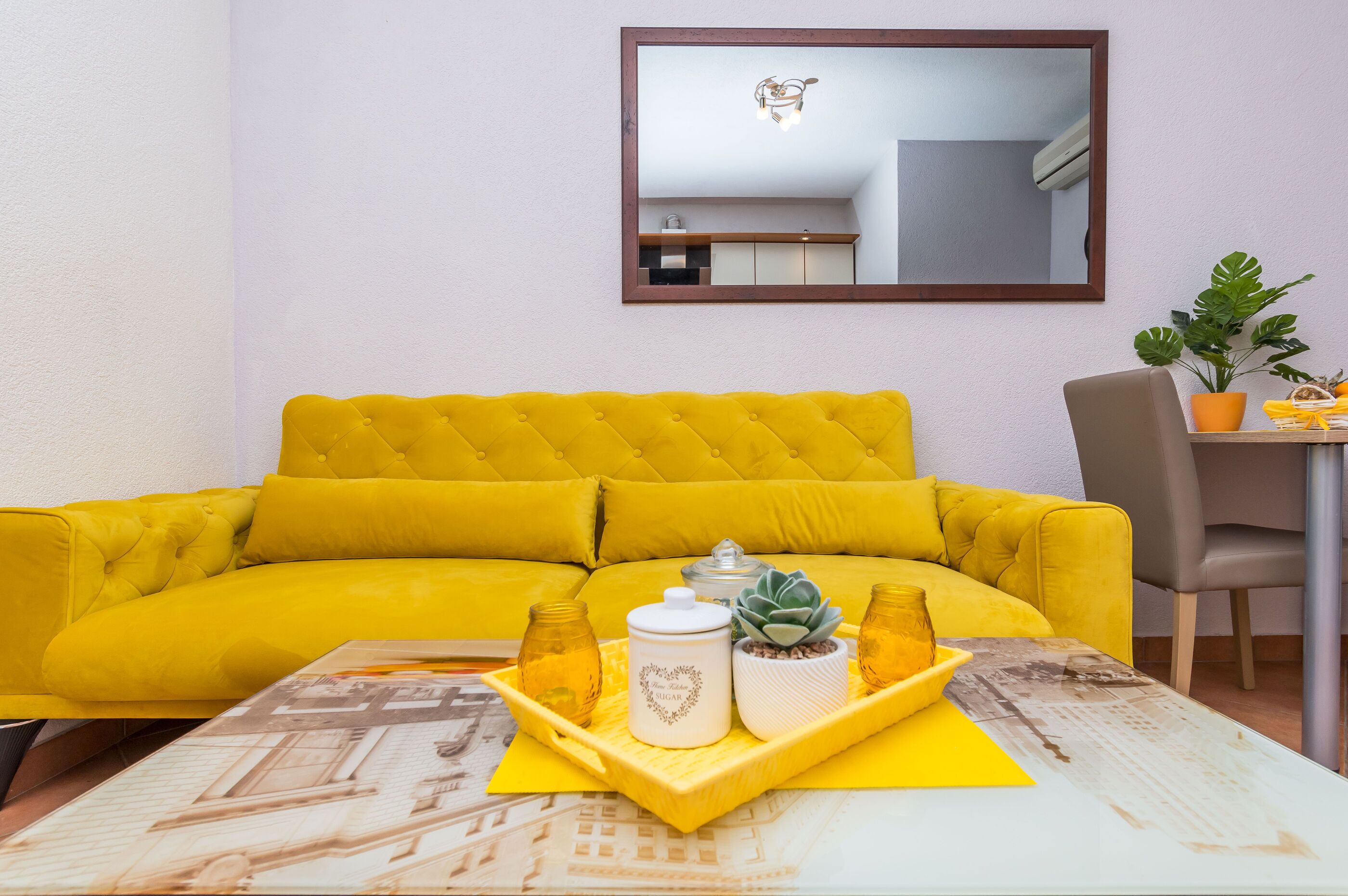 A bright yellow sofa with a tufted backrest, a small table with a tray holding a jar, a potted plant, and a white container with a heart design.