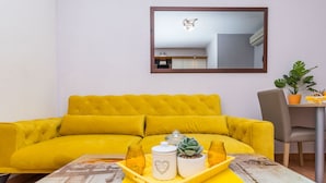 A bright yellow sofa with a tufted backrest, a small table with a tray holding a jar, a potted plant, and a white container with a heart design. - Apartment Piveta (Okrug Gornji)