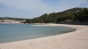 A beach with clear water, a pebbly shore, and a forested hill in the background. - Apartment Lidija (Tisno)