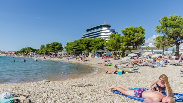 A pebbled beach with people sunbathing and swimming, a modern building in the background, and trees providing shade.