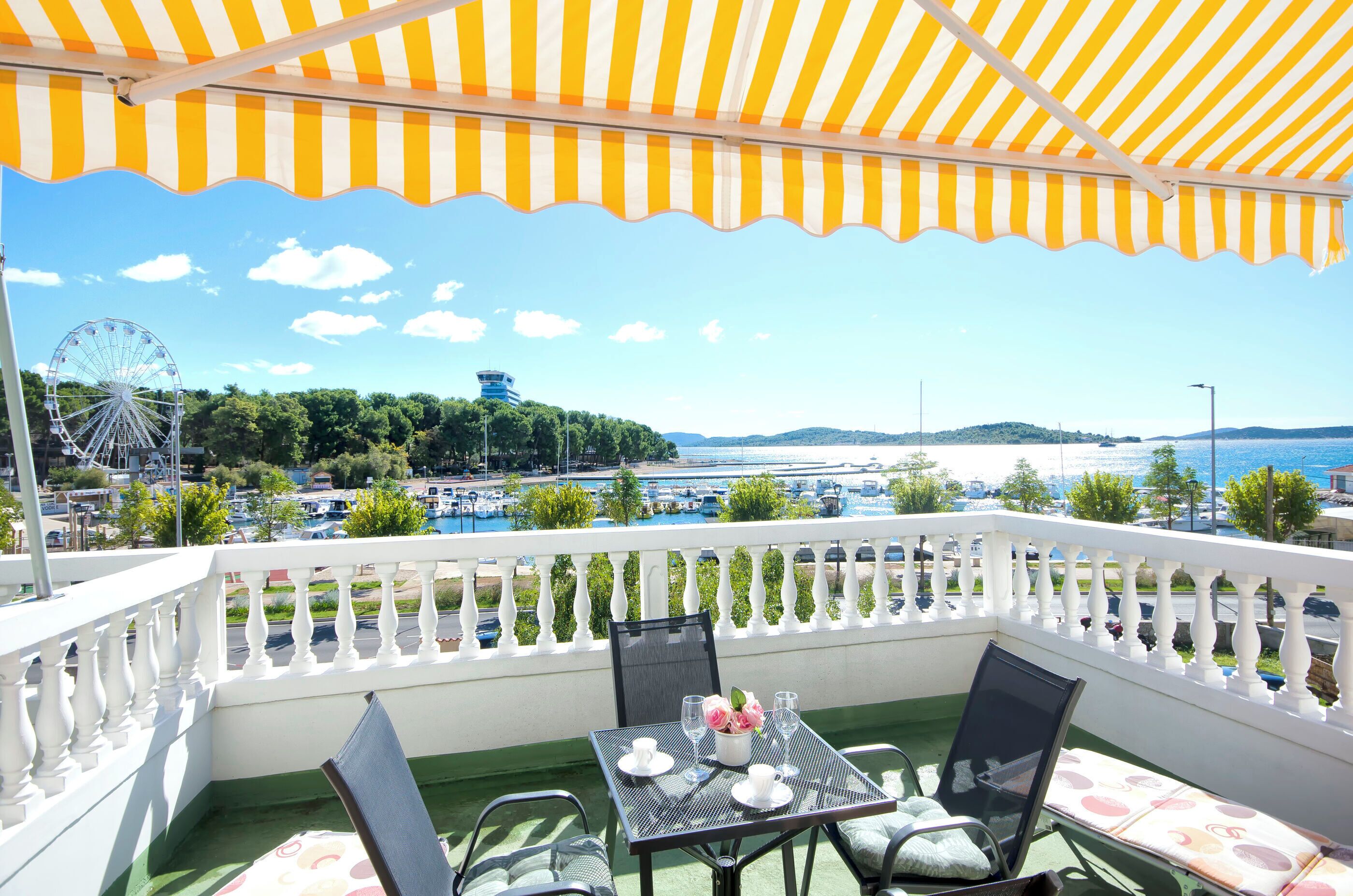 A balcony with a table set for two, overlooking a scenic view of a body of water, a Ferris wheel, and distant hills.