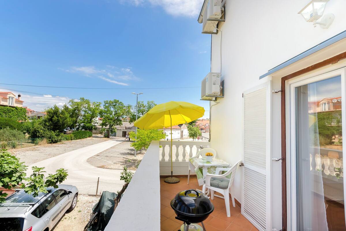 A balcony with a yellow umbrella, a grill, and a table with chairs.