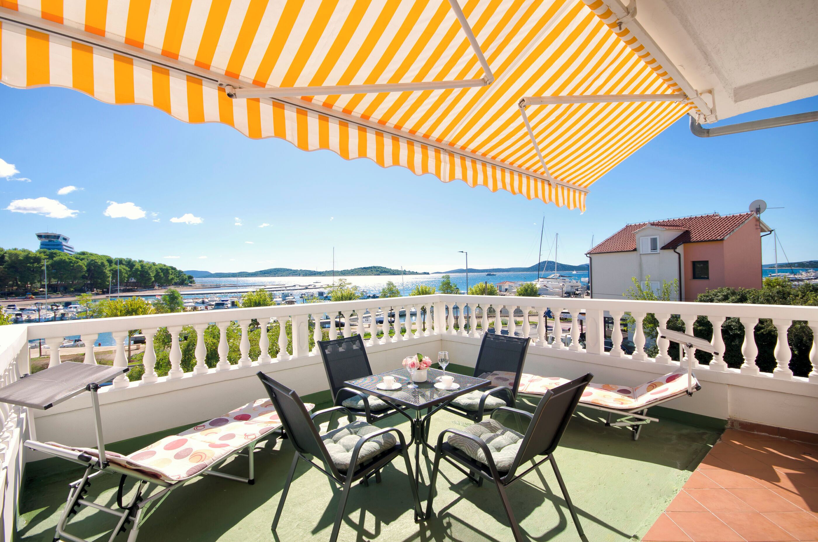 En balkon med en stribet markise, et bord med stole og udsigt til havet og bygninger.
