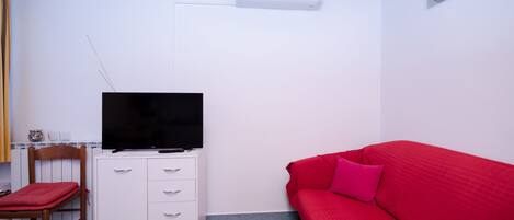 A room with a TV on a cabinet, a red couch, a wooden chair, and an air conditioning unit on the wall.