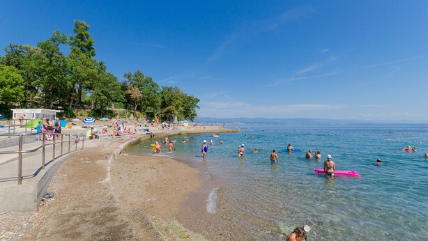 Une plage avec une eau claire, des gens qui se baignent et prennent des bains de soleil, et quelques parasols.