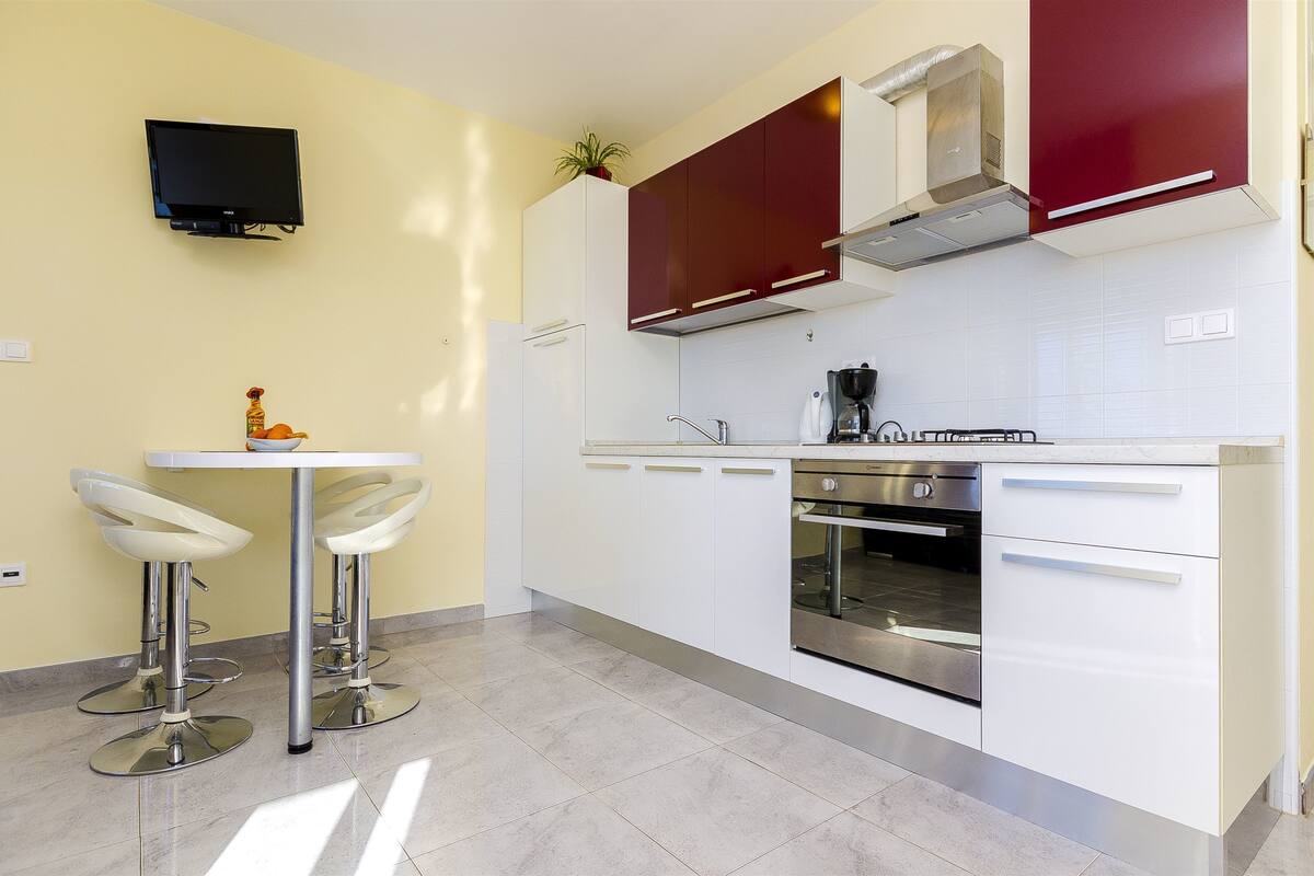 A modern kitchen with white cabinets, a stainless steel oven, and a flat-screen TV mounted on the wall.