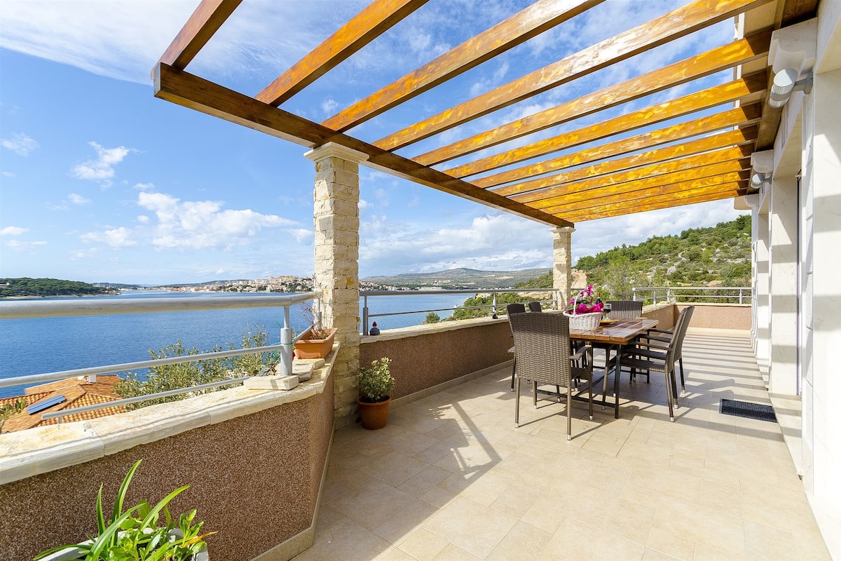 A terrace with a table and chairs, overlooking a body of water and a distant landscape.