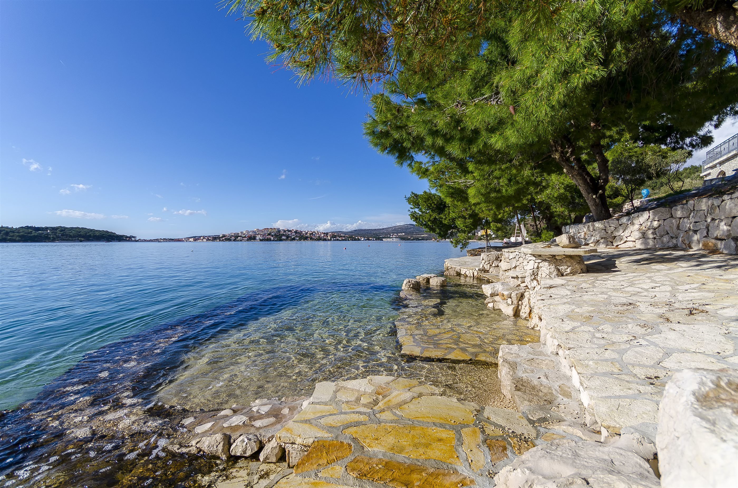 A stone waterfront with clear water, a stone wall, and a view of a distant town.