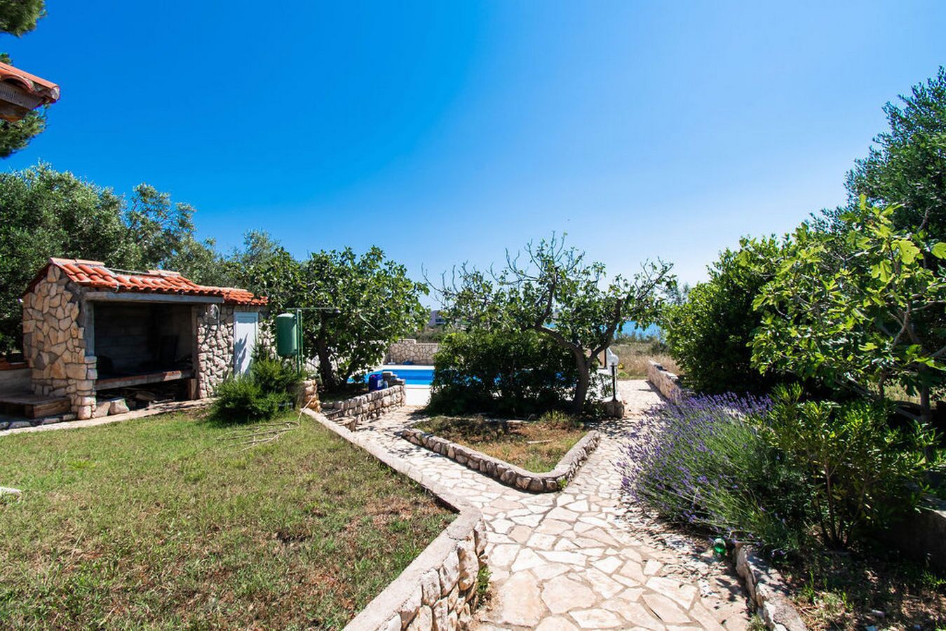 A stone-built outdoor kitchen with a tiled roof, a stone pathway leading to a pool, and surrounding greenery.