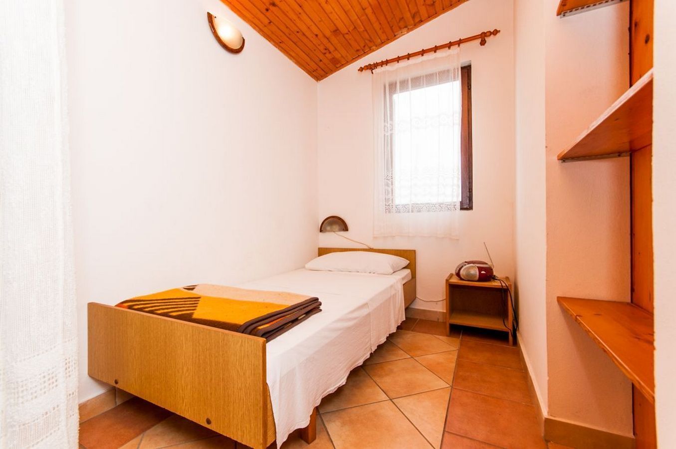 Une petite chambre avec un lit simple, une tête de lit en bois, une table de chevet avec une lampe et une fenêtre ornée d’un rideau léger.