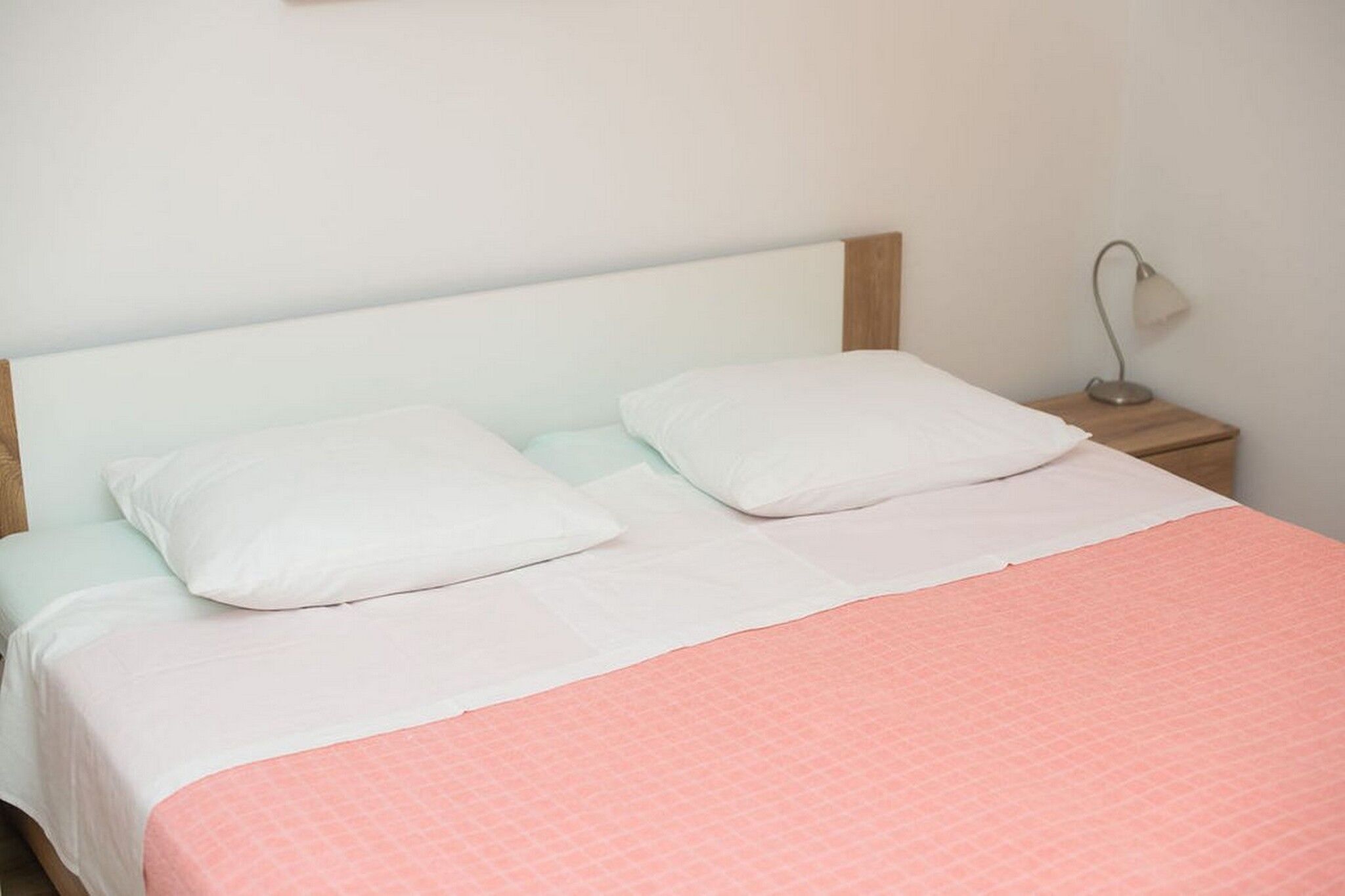 Un lit avec une tête de lit blanche, deux oreillers blancs, une couvre-lit rose et une table de chevet avec une lampe.