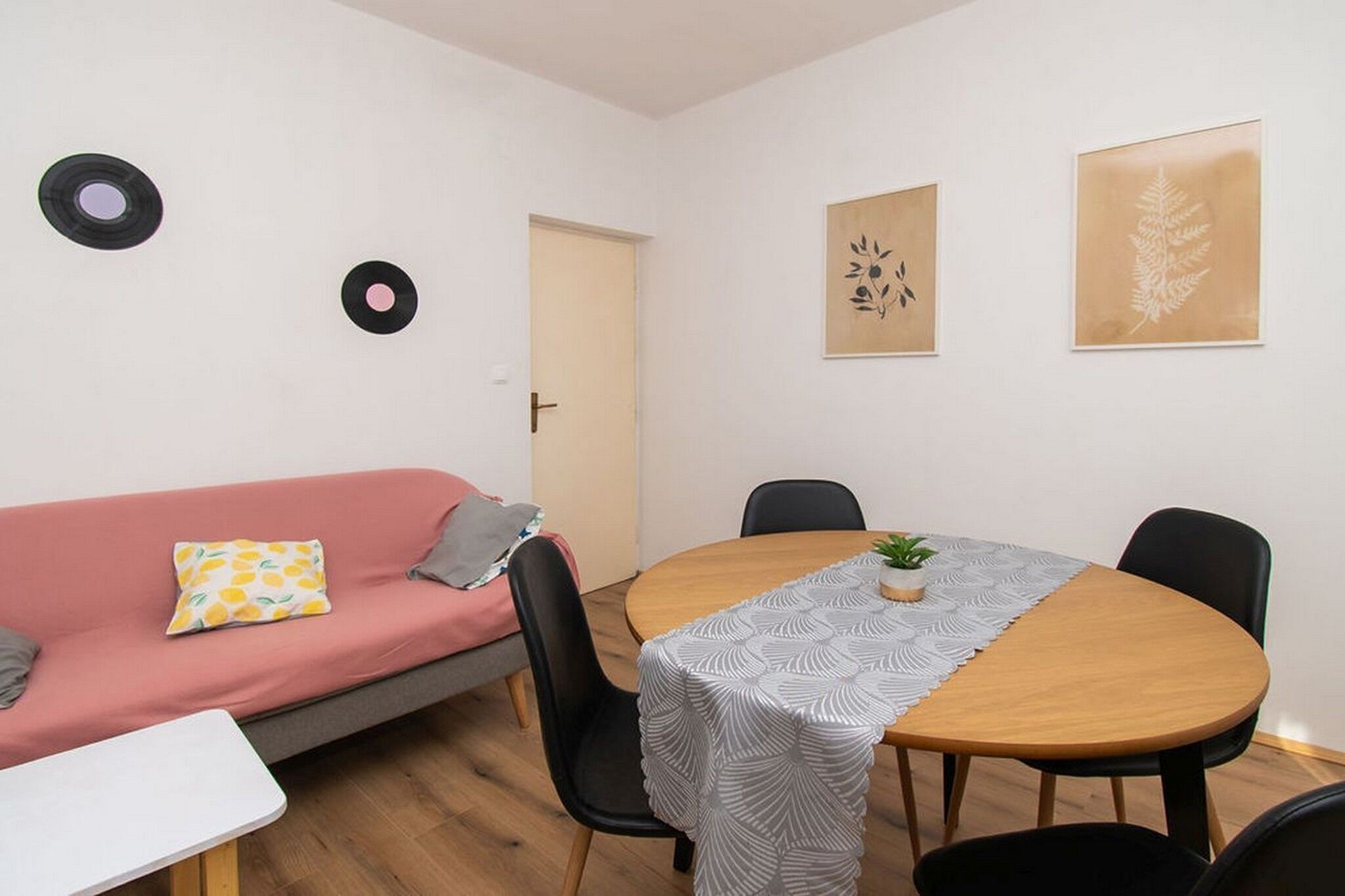 A small, modern room with a round wooden table, black chairs, a pink sofa, and a white table. There are two vinyl records on the wall and two framed botanical prints.