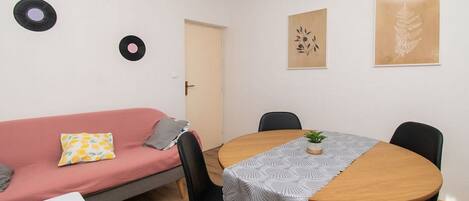 A small, modern room with a round wooden table, black chairs, a pink sofa, and a white table. There are two vinyl records on the wall and two framed botanical prints.