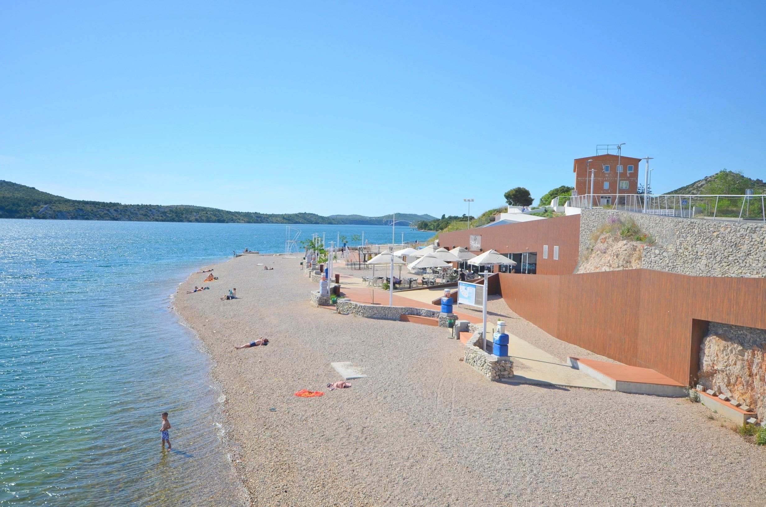 A beach with a pebble shore, a building with a red facade, and a clear blue sky.