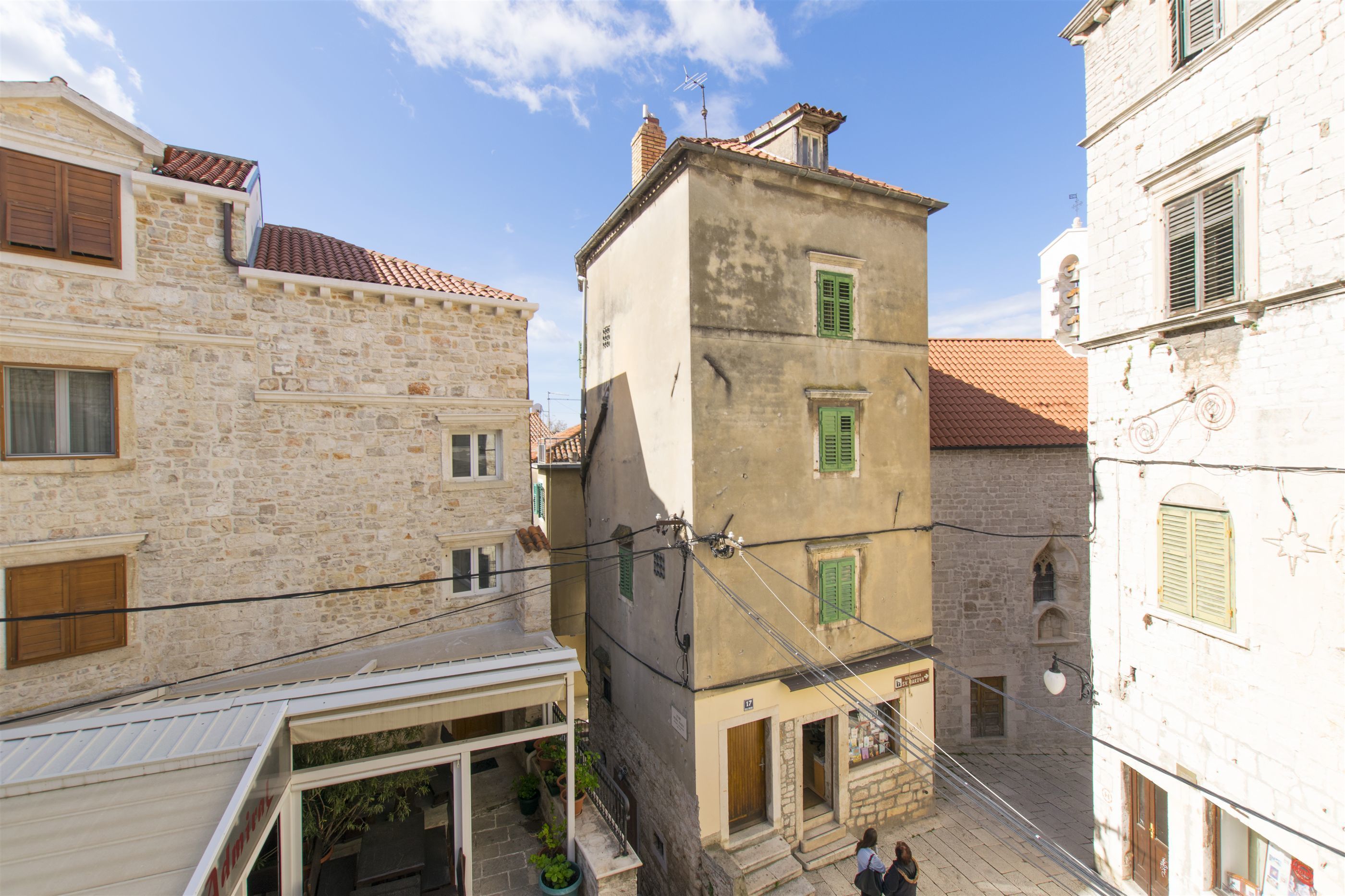 Una calle estrecha con edificios de piedra, una tienda visible a la izquierda y una escalera que sube a un piso superior.