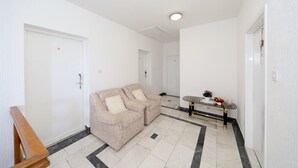 A clean, white-walled room with a beige sofa, a small table with a vase and fruit, and a wooden bench. - Apartments Family Gligora (Mandre)