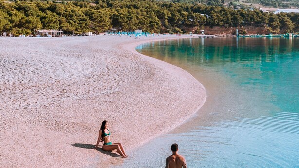 Uma praia com águas turquesa cristalinas, faixa de areia e montanhas ao fundo.