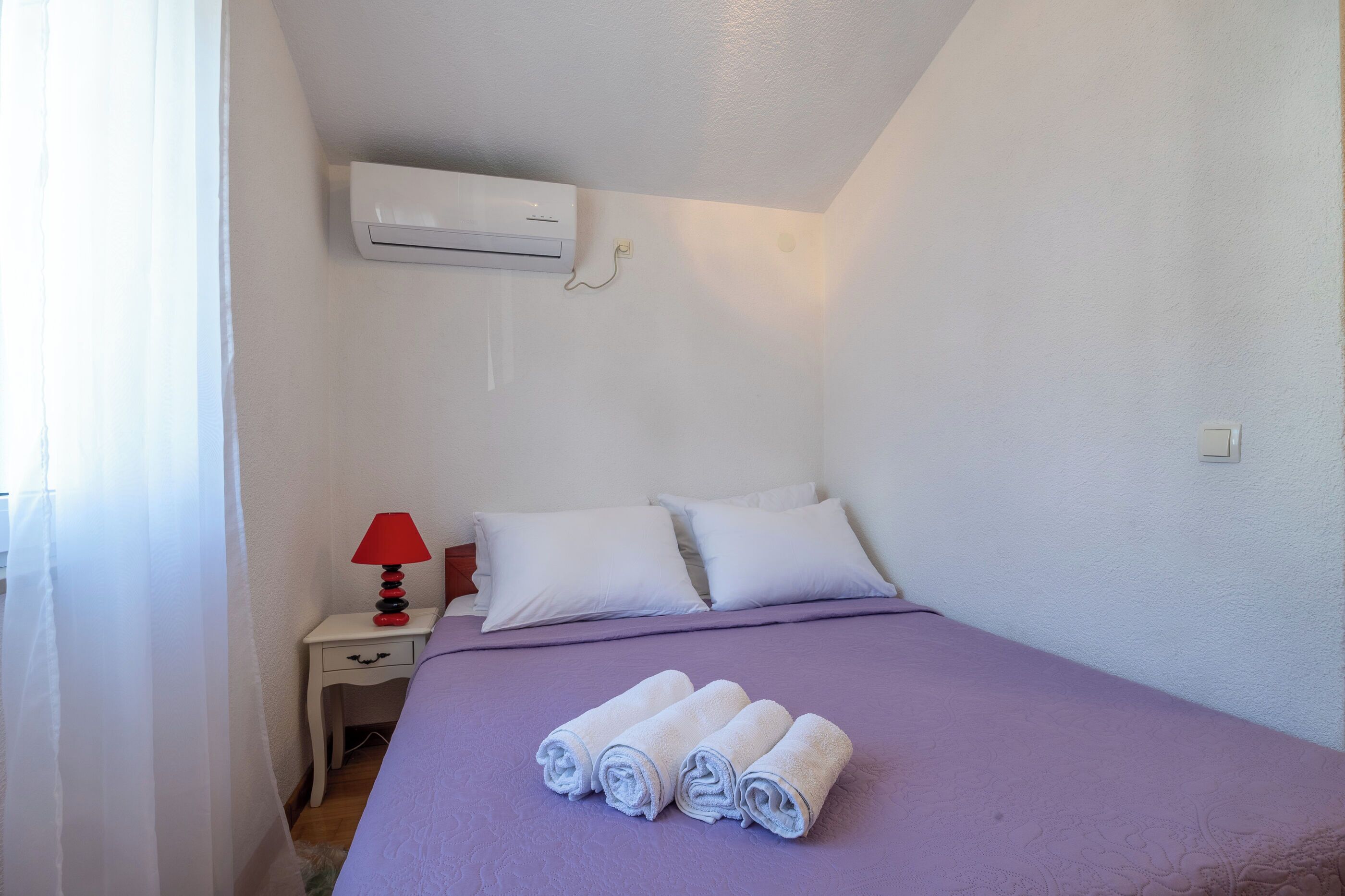 A bedroom with a bed, white pillows, a red lamp, a white nightstand, and a rolled-up towel on the bed.