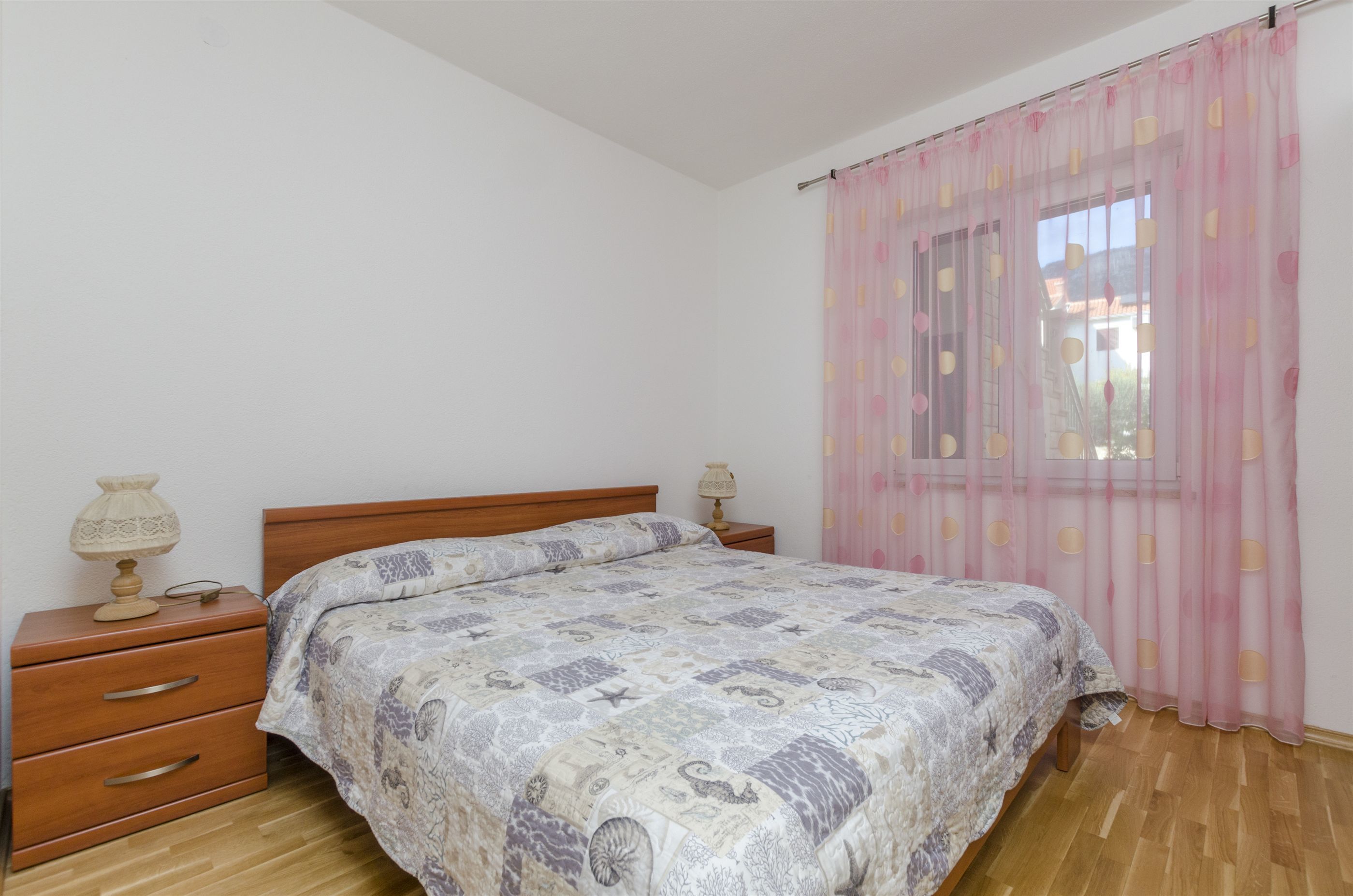 A bedroom with a wooden bed, two bedside tables with lamps, a window with pink curtains, and a wooden floor.