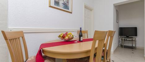 A dining area with a round table, chairs, a fruit bowl, a bottle, and a television in the background.