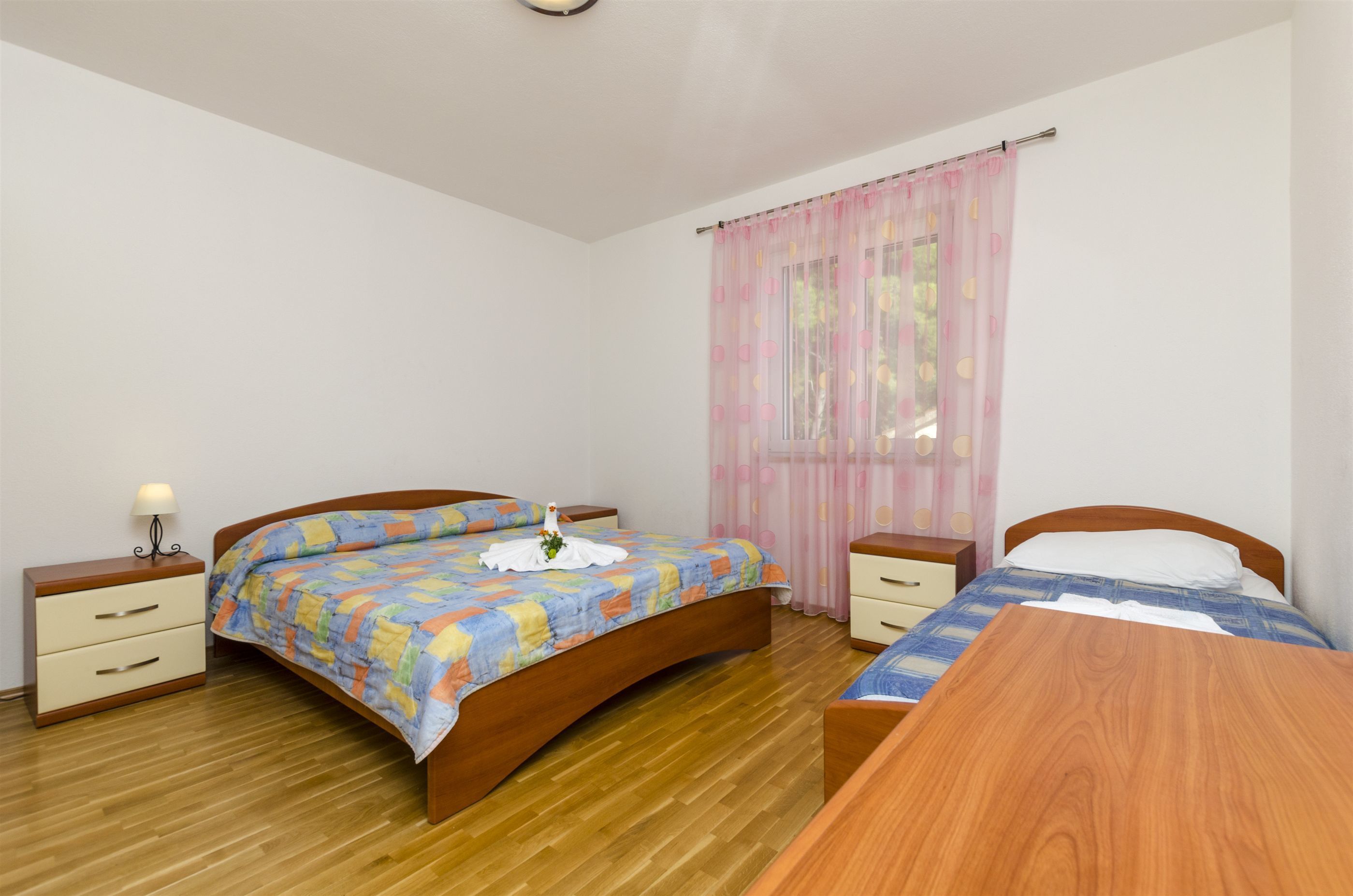 A bedroom with a wooden bed, a bedside table, a nightstand, and a window with sheer curtains.