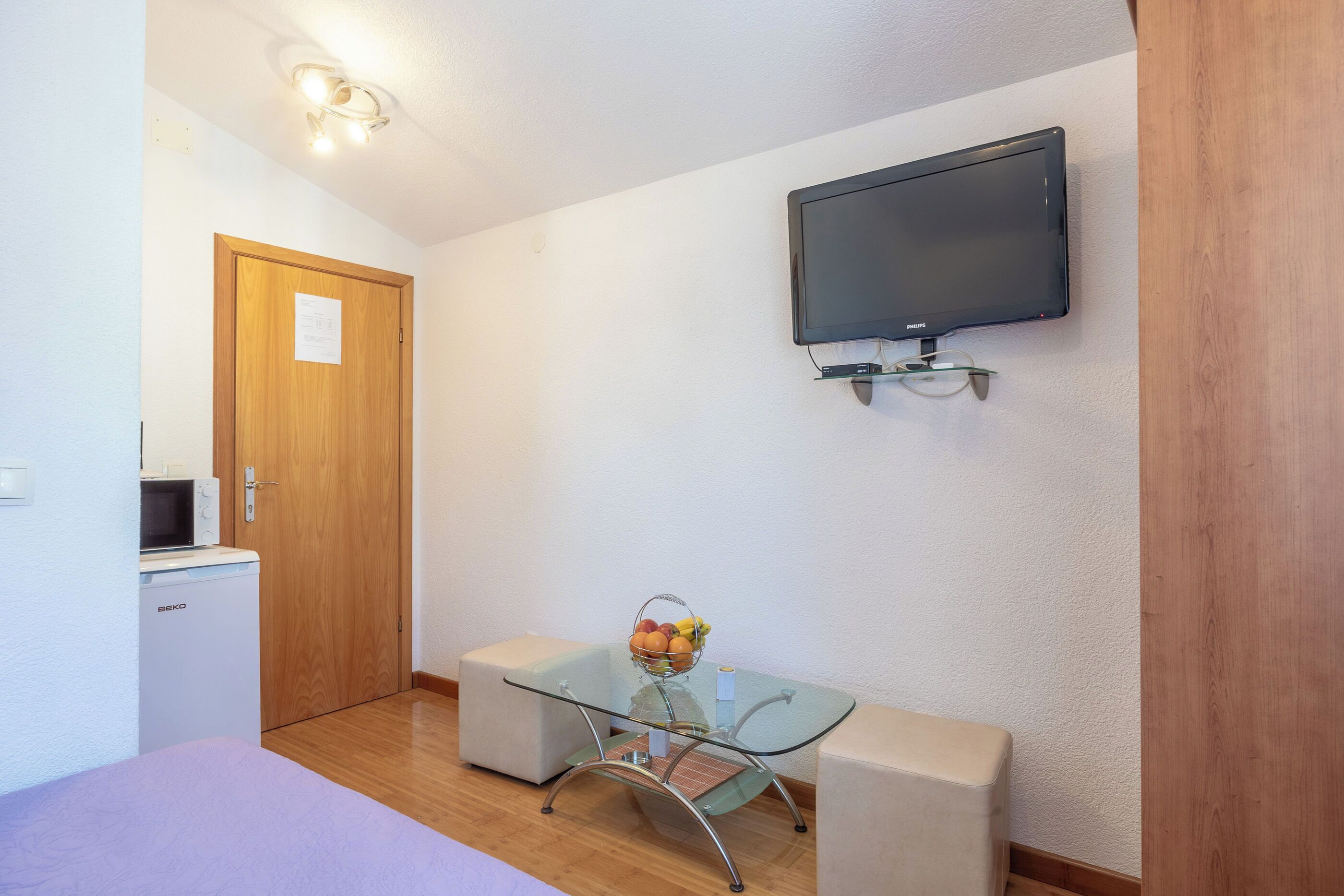 A compact room with a bed, a small kitchenette, a glass table with chairs, a flat-screen TV, and a wooden door.