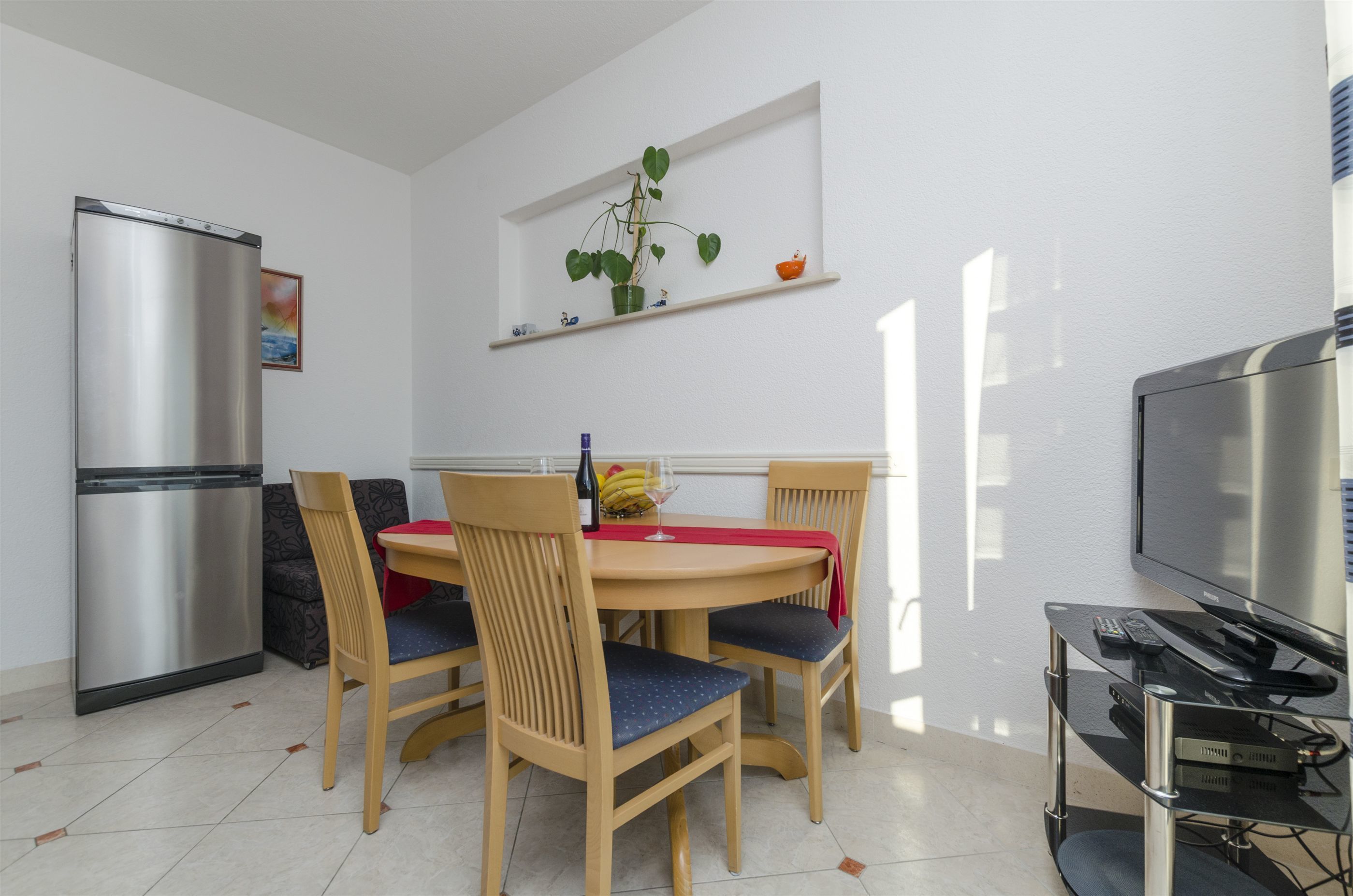 A dining area with a round table, four chairs, a refrigerator, a TV, and a shelf with a plant.