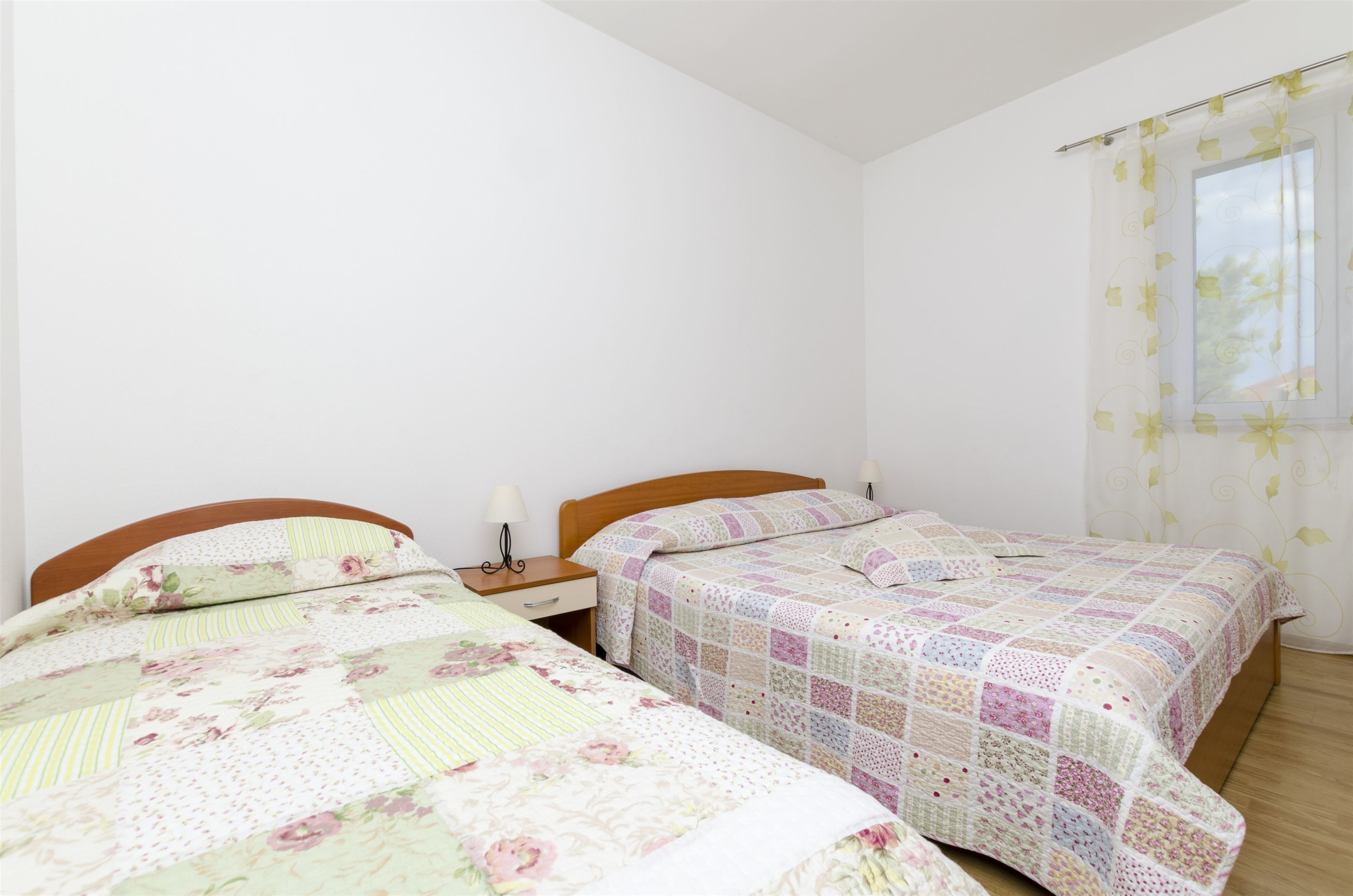 A bedroom with two single beds, a wooden headboard, a nightstand with a lamp, and a window with floral curtains.