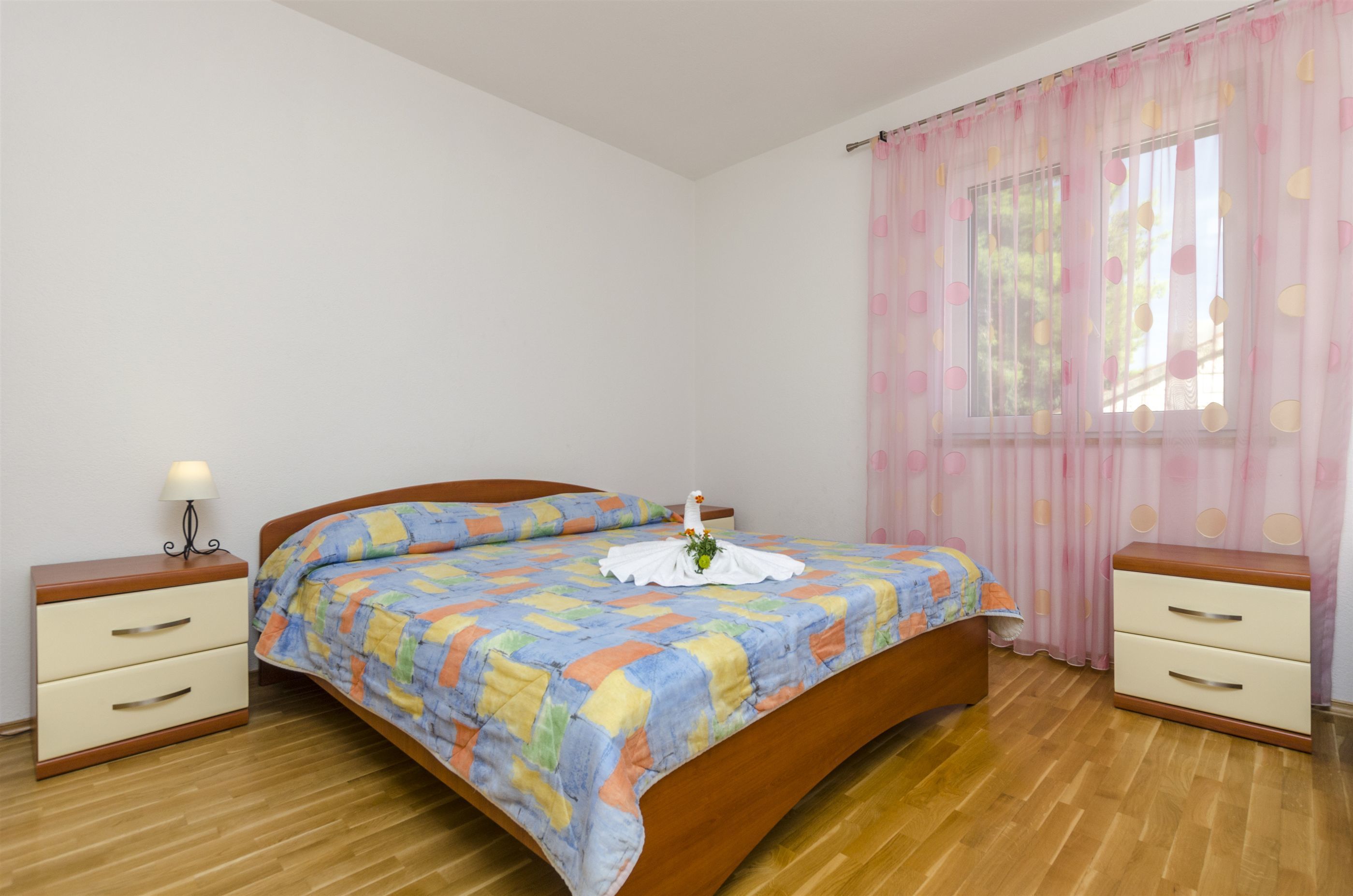 A neatly made bed with a colorful quilt, a bedside table with a lamp, and a window with pink curtains.