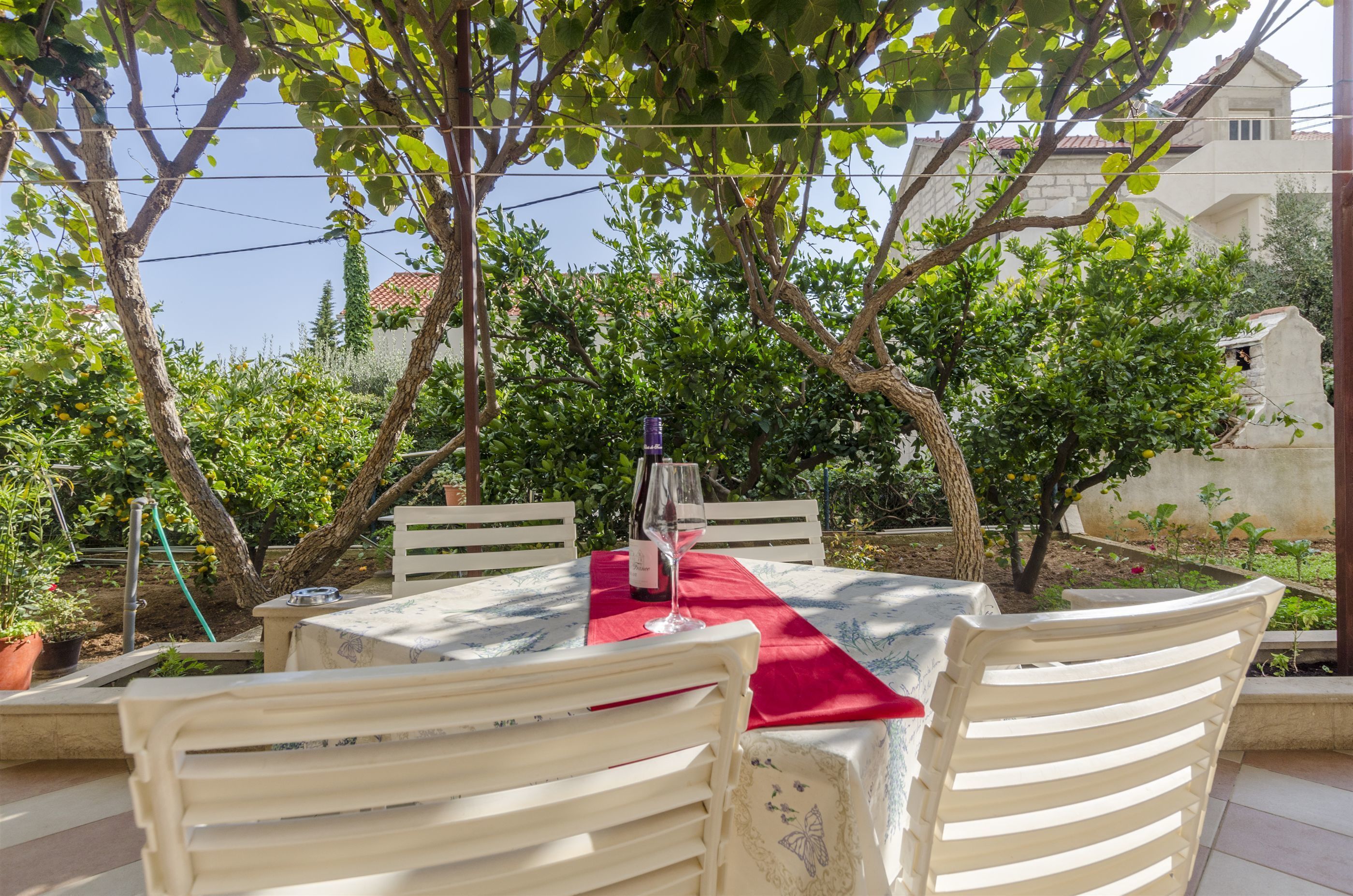 An outdoor dining area with a table set for two, surrounded by white chairs, under a tree.