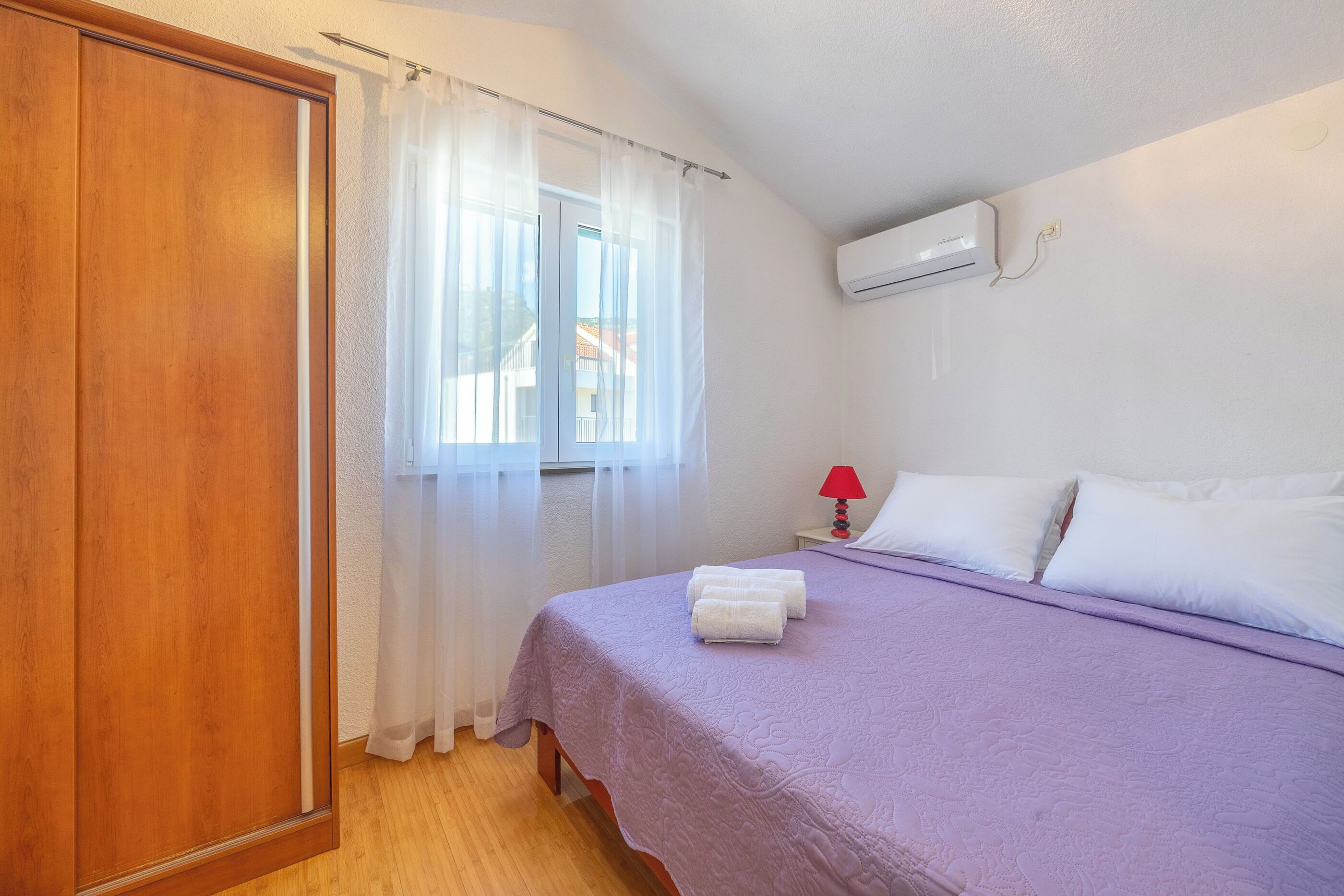 A bedroom with a bed, white curtains, a red lamp, and an air conditioning unit.