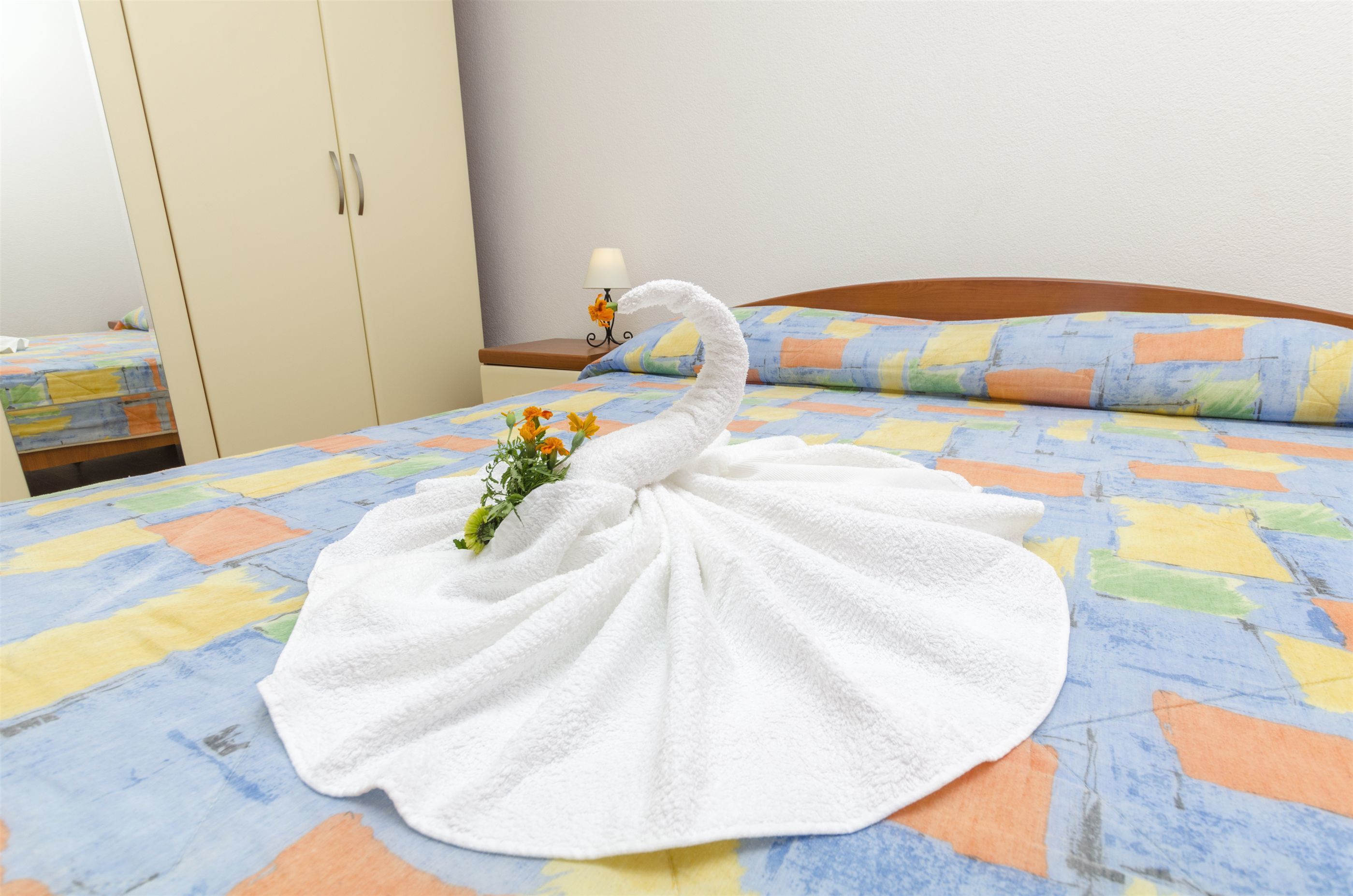 Un lit bien fait, recouvert d’une courtepointe aux couleurs vives, une serviette blanche en forme de cygne et une petite plante posée sur le lit.