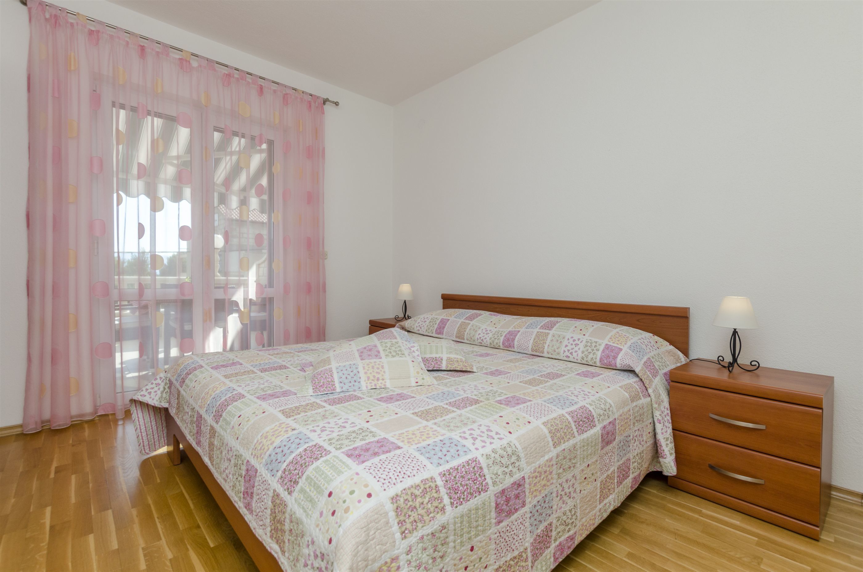 Ein Schlafzimmer mit einem hölzernen Bett, einem Nachttisch mit Lampe und einem Fenster mit rosa Vorhängen.