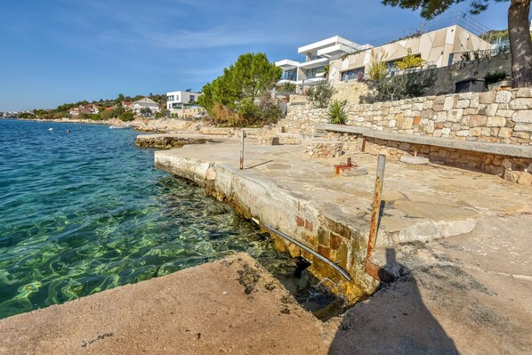 Zona costera con aguas transparentes, un muelle de piedra y edificios modernos con balcones con vista al mar.